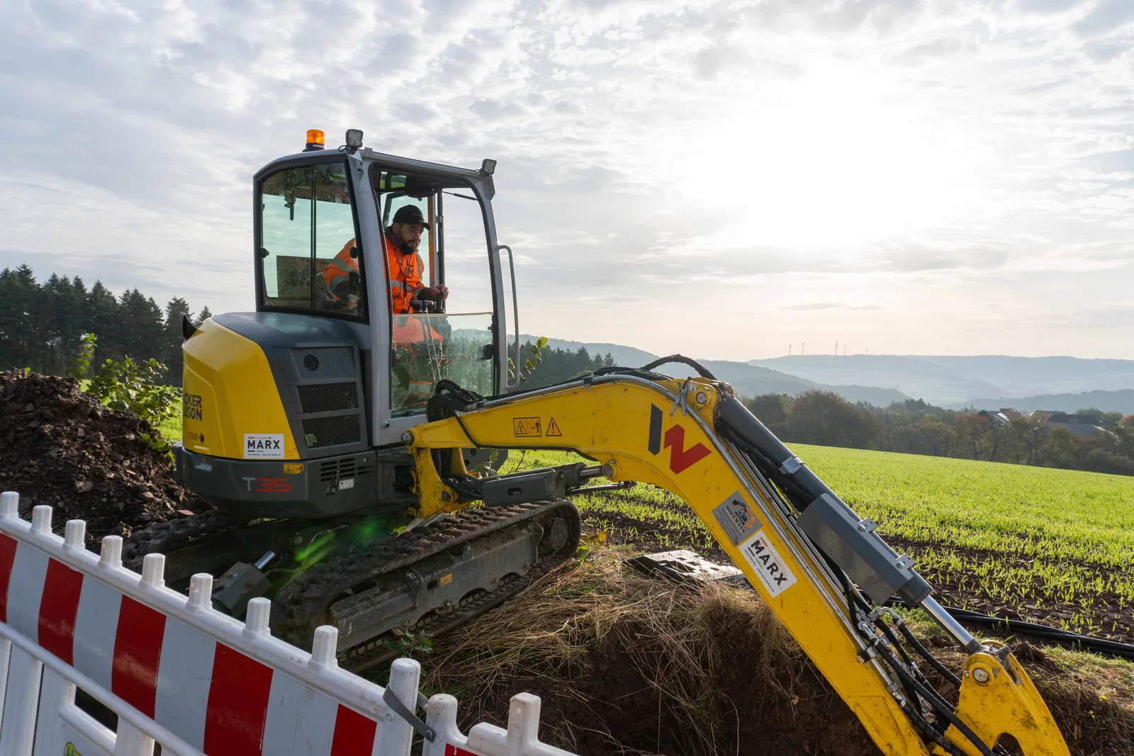 Minibagger hebt auf einer Baustelle von MARX Tiefbau eine Grube aus, im Hintergrund Felder