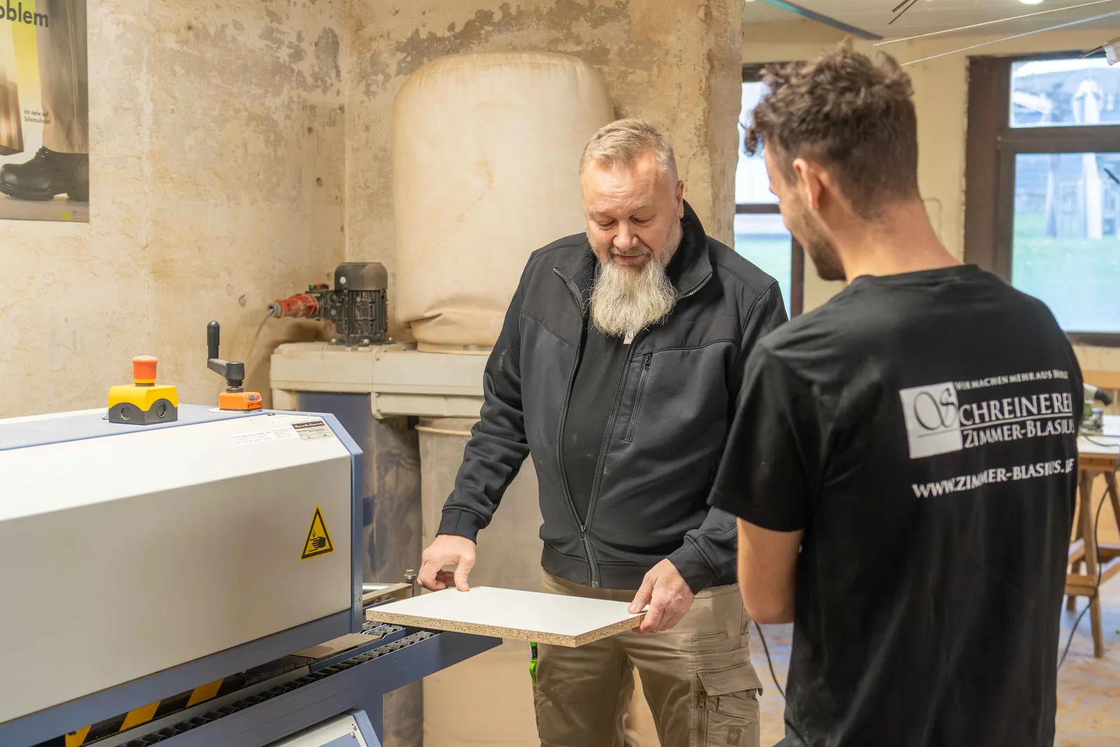 Schreinermeister erklärt einem jungen Mitarbeiter ein Werkstück an der Kantenmaschine der Schreinerei Zimmer-Blasius