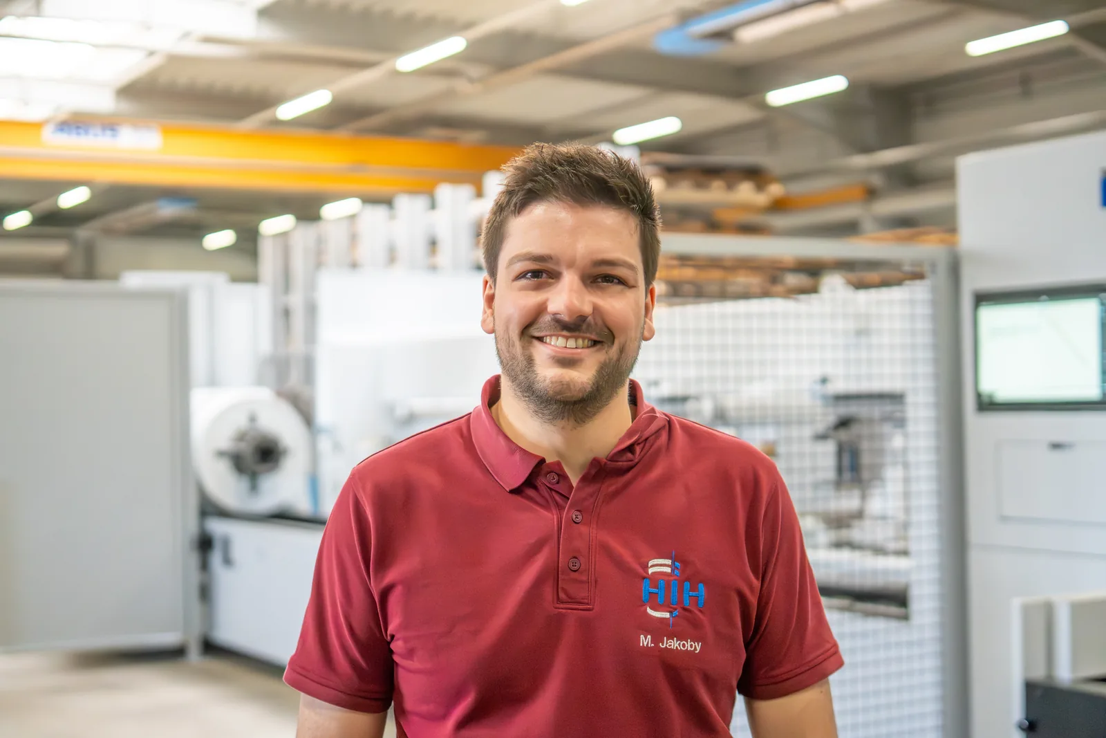 Lächelnder Mitarbeiter im roten Poloshirt vor Maschinen in der Werkhalle von Hand in Hand Industriemontage