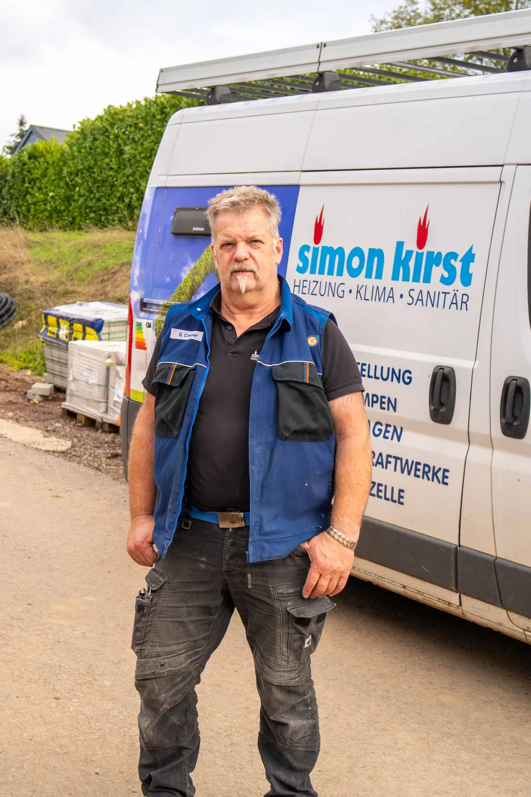 Monteur der Simon Kirst GmbH in Arbeitsweste steht vor dem Firmenwagen mit Logo auf der Baustelle