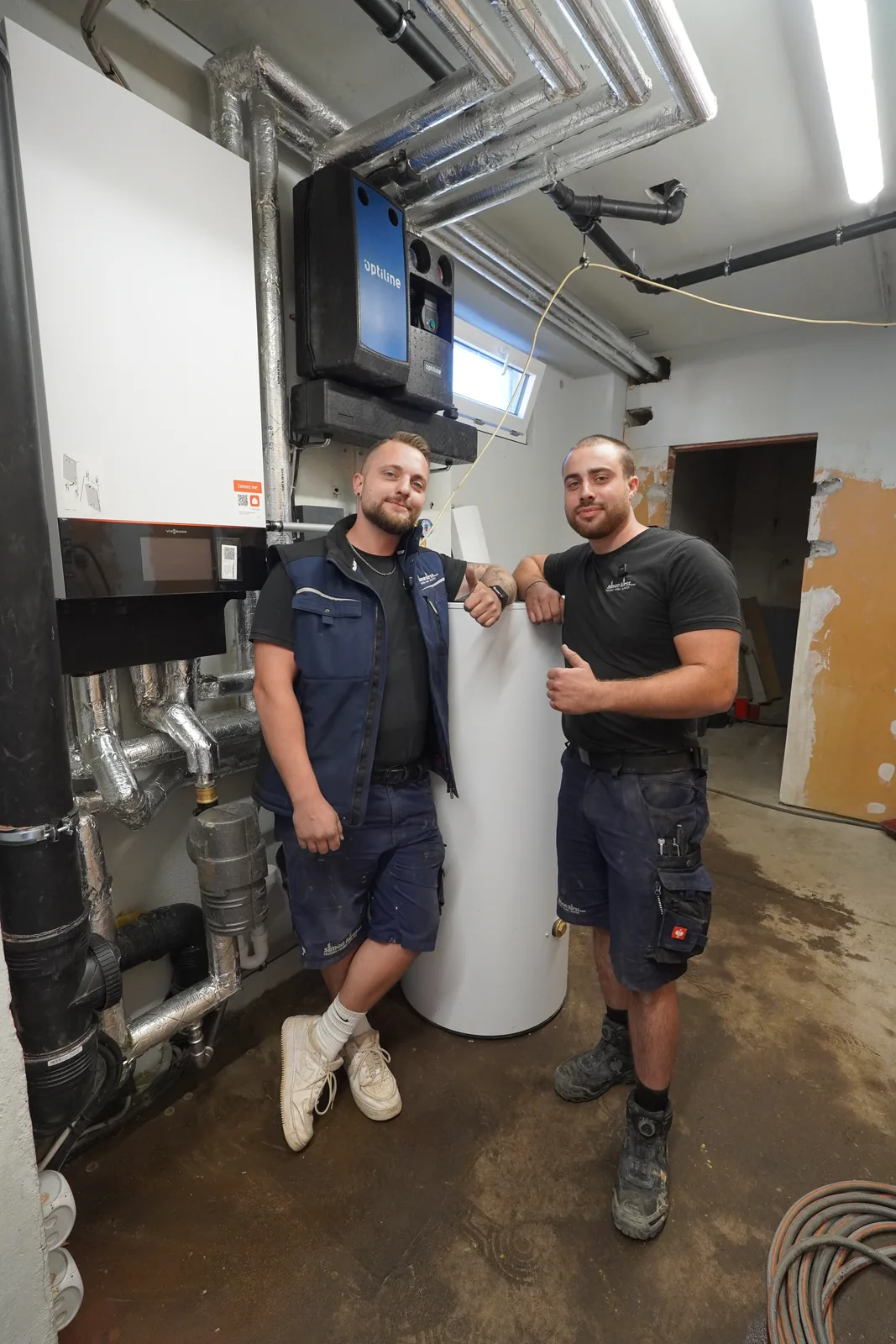 Zwei Monteure der Simon Kirst GmbH lehnen gut gelaunt an einem Speicher im Heizungsraum, einer zeigt den Daumen nach oben