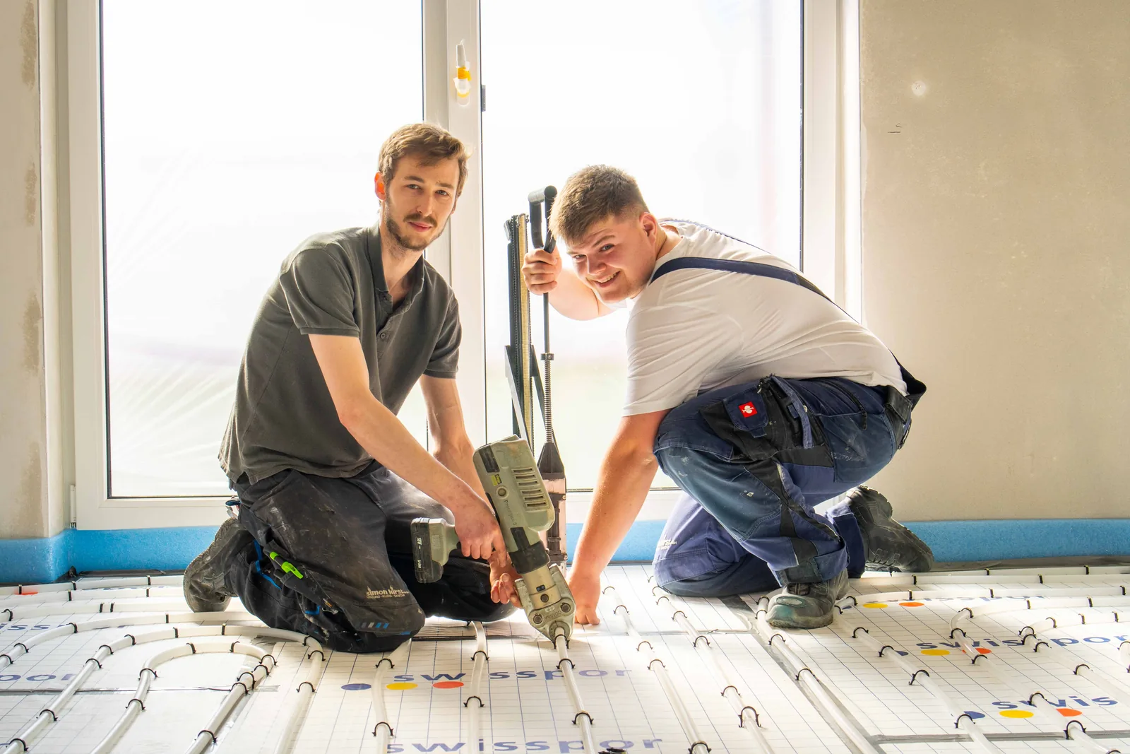 Zwei Monteure der Simon Kirst GmbH knien lächelnd auf der Baustelle und verlegen eine Fußbodenheizung mit dem Tackergerät