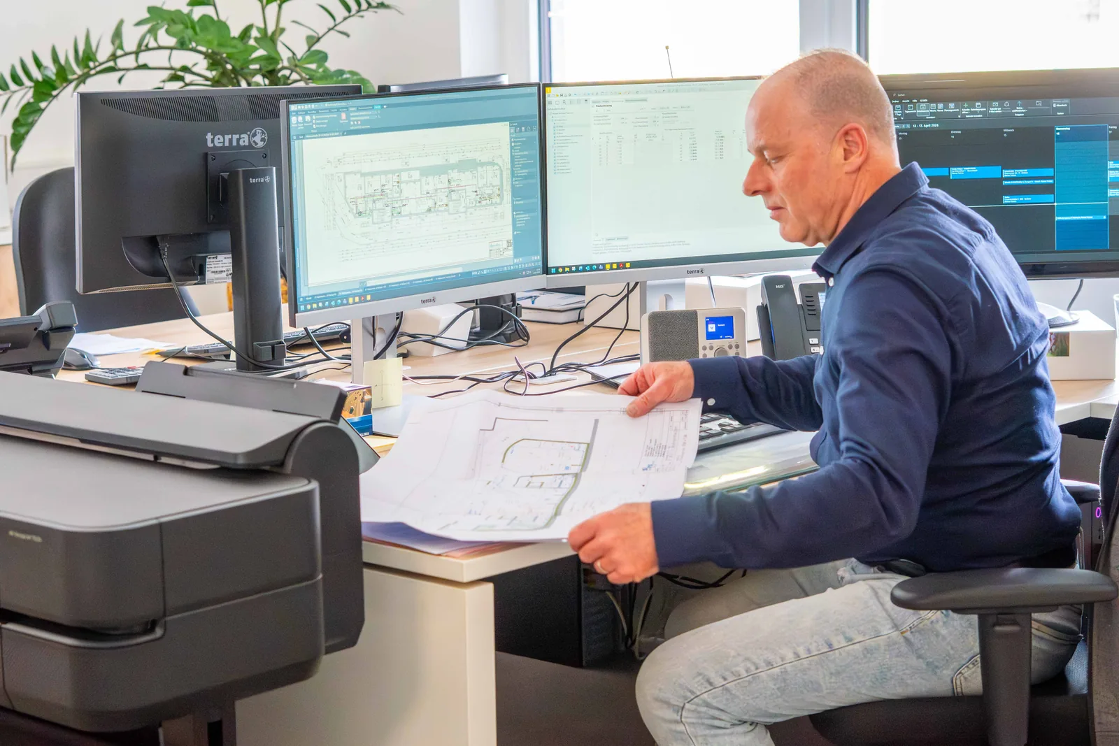 Am Schreibtisch der Simon Kirst GmbH wird ein Bauplan geprüft, daneben Bildschirme mit Planungssoftware