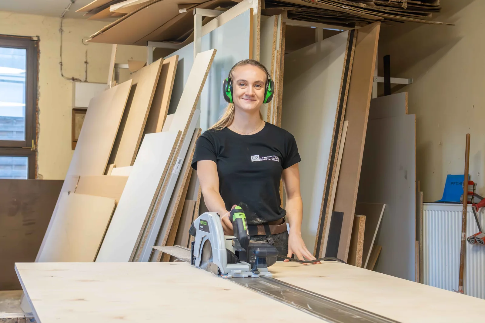 Junge Schreinerin mit Gehörschutz sägt eine Holzplatte in der Werkstatt der Schreinerei Zimmer-Blasius
